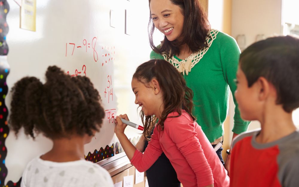 Teacher smiling with 3 students at whiteboard, one writing